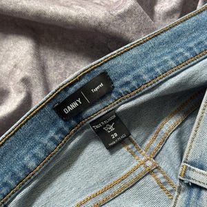May include: Close-up of light blue denim jeans with visible stitching. The jeans have a "DANNY" label and a "True Religion" tag indicating size 29, made in Mexico. The jeans are tapered.