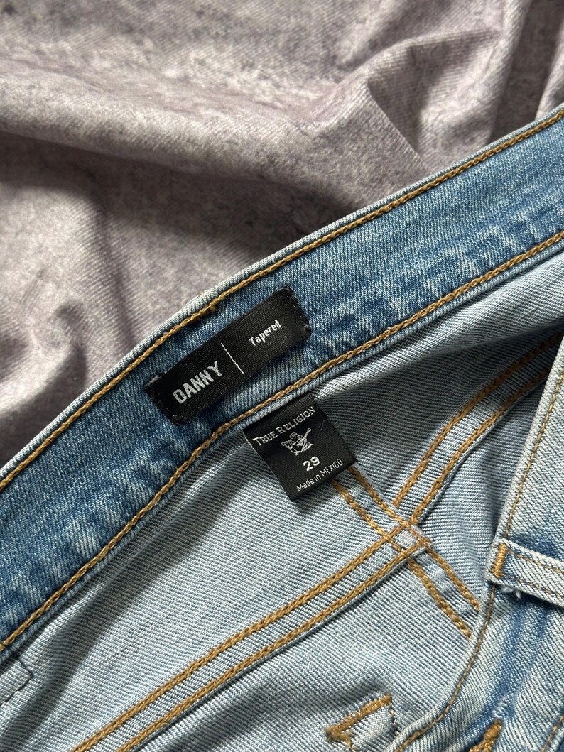 May include: Close-up of light blue denim jeans with visible stitching. The jeans have a "DANNY" label and a "True Religion" tag indicating size 29, made in Mexico. The jeans are tapered.
