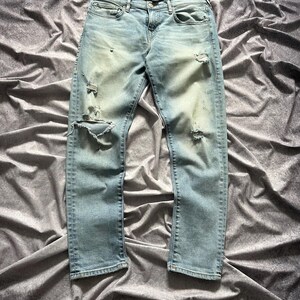 May include: Light blue denim jeans with distressed details, including rips on the knees and thighs. The jeans have a classic five-pocket design and a button and zipper closure. The denim has a faded wash.