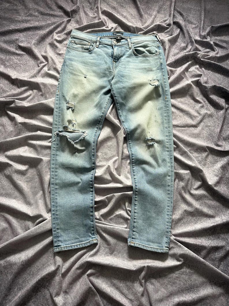 May include: Light blue denim jeans with distressed details, including rips on the knees and thighs. The jeans have a classic five-pocket design and a button and zipper closure. The denim has a faded wash.