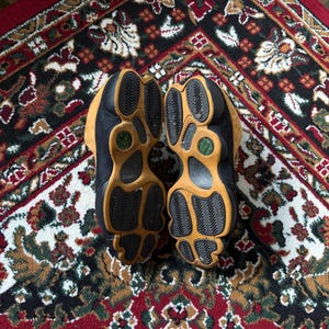 Op de afbeelding: Paar zwarte en bruine sneakers met een textuurzool, rustend op een patroon rood, zwart en bruin tapijt. De zolen zijn lichtbruin met zwart profiel en een groene accentkleur.