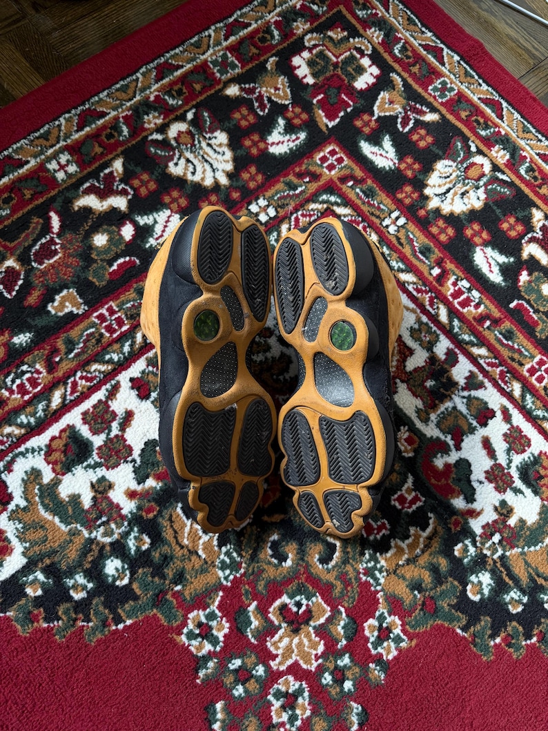 Op de afbeelding: Paar zwarte en bruine sneakers met een textuurzool, rustend op een patroon rood, zwart en bruin tapijt. De zolen zijn lichtbruin met zwart profiel en een groene accentkleur.