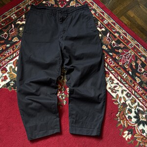 May include: Black cropped trousers with a relaxed fit. The trousers are made of a solid, dark fabric and have a button and zip closure. The trousers are displayed on a patterned rug with red and brown tones.