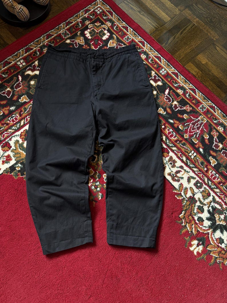 May include: Black cropped trousers with a relaxed fit. The trousers are made of a solid, dark fabric and have a button and zip closure. The trousers are displayed on a patterned rug with red and brown tones.