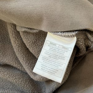 May include: Close-up of a taupe-colored hoodie interior with a white tag. The tag displays the garment's composition: 80% cotton, 20% polyester, and 100% cotton hood lining. The tag also indicates the item was made in Indonesia.