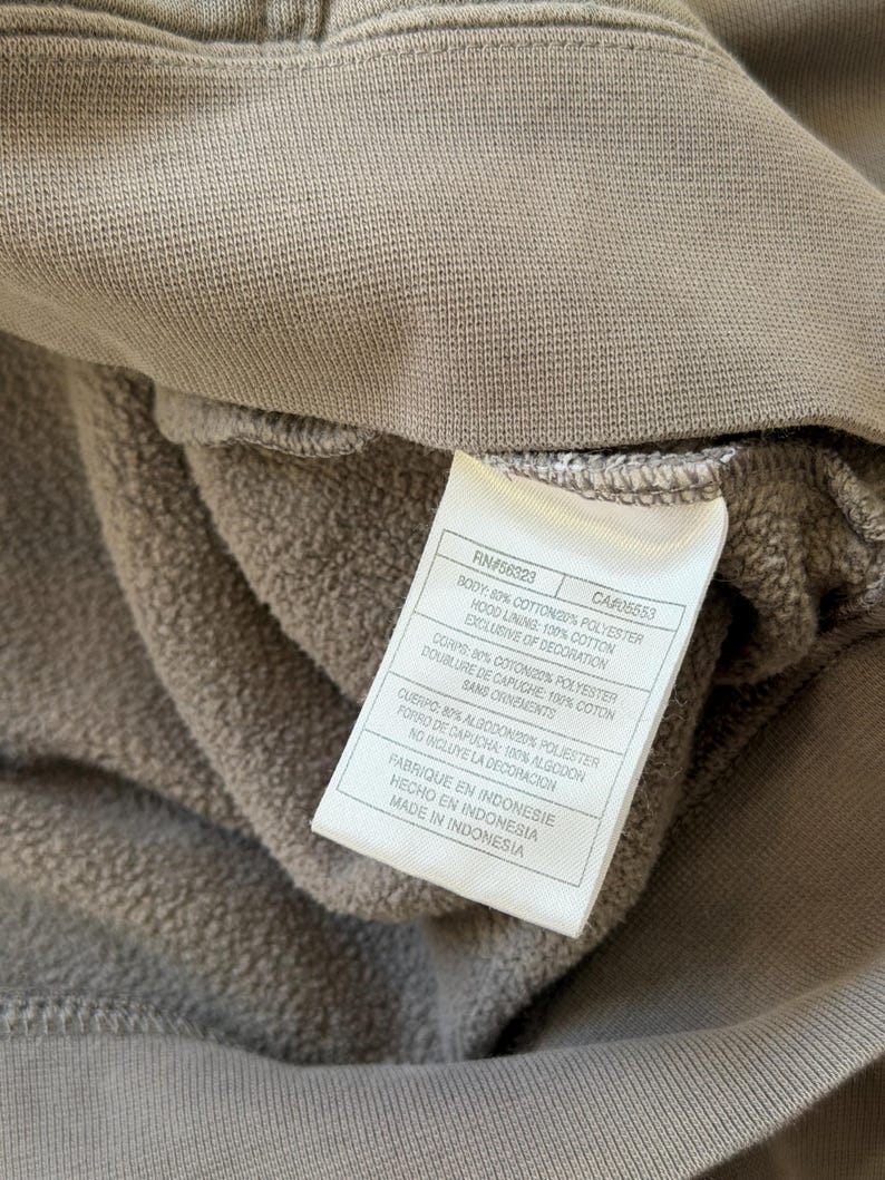 May include: Close-up of a taupe-colored hoodie interior with a white tag. The tag displays the garment's composition: 80% cotton, 20% polyester, and 100% cotton hood lining. The tag also indicates the item was made in Indonesia.