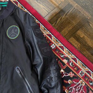 Może przedstawiać: Czarna kurtka bomberka z okrągłą naszywką z numerem 23 i napisem "Jordan". Kurtka ma kieszeń zapinaną na zamek i naszywki na rękawie ze szczeg&oacute;łami w postaci gwiazd i tekstu. Kurtka leży na czerwono-czarnym dywanie.