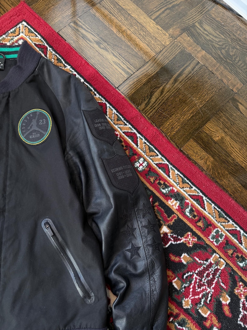 Może przedstawiać: Czarna kurtka bomberka z okrągłą naszywką z numerem 23 i napisem "Jordan". Kurtka ma kieszeń zapinaną na zamek i naszywki na rękawie ze szczeg&oacute;łami w postaci gwiazd i tekstu. Kurtka leży na czerwono-czarnym dywanie.