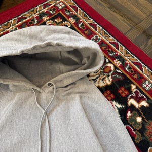 Pu&ograve; includere: Una felpa con cappuccio grigio chiaro con coulisse. La felpa con cappuccio &egrave; realizzata in un tessuto morbido e strutturato. Lo sfondo include un tappeto rosso con un motivo dettagliato e pavimenti in legno.