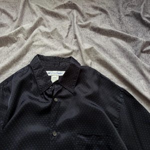 May include: Black button-down shirt with a subtle polka dot pattern. The shirt has a classic collar, a chest pocket, and a buttoned front. The fabric has a slight sheen.