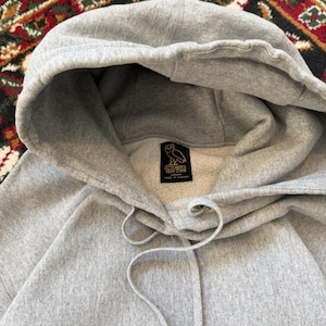 Pu&ograve; includere: Una felpa con cappuccio grigio chiaro con coulisse e cappuccio. La felpa ha un'etichetta con un logo e del testo. Il tessuto sembra essere un materiale morbido e di peso medio. Lo sfondo &egrave; un tappeto fantasia.