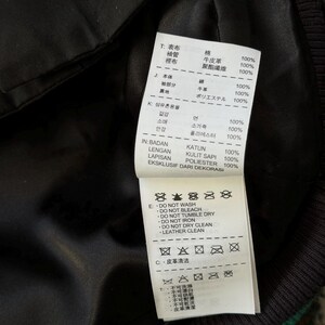 Może przedstawiać: Zbliżenie na czarną podszewkę kurtki z białą metką. Metka zawiera tekst w wielu językach, w tym japońskim i angielskim, szczeg&oacute;łowo opisujący skład tkaniny i instrukcje pielęgnacji. Kurtka ma zielony pasek.