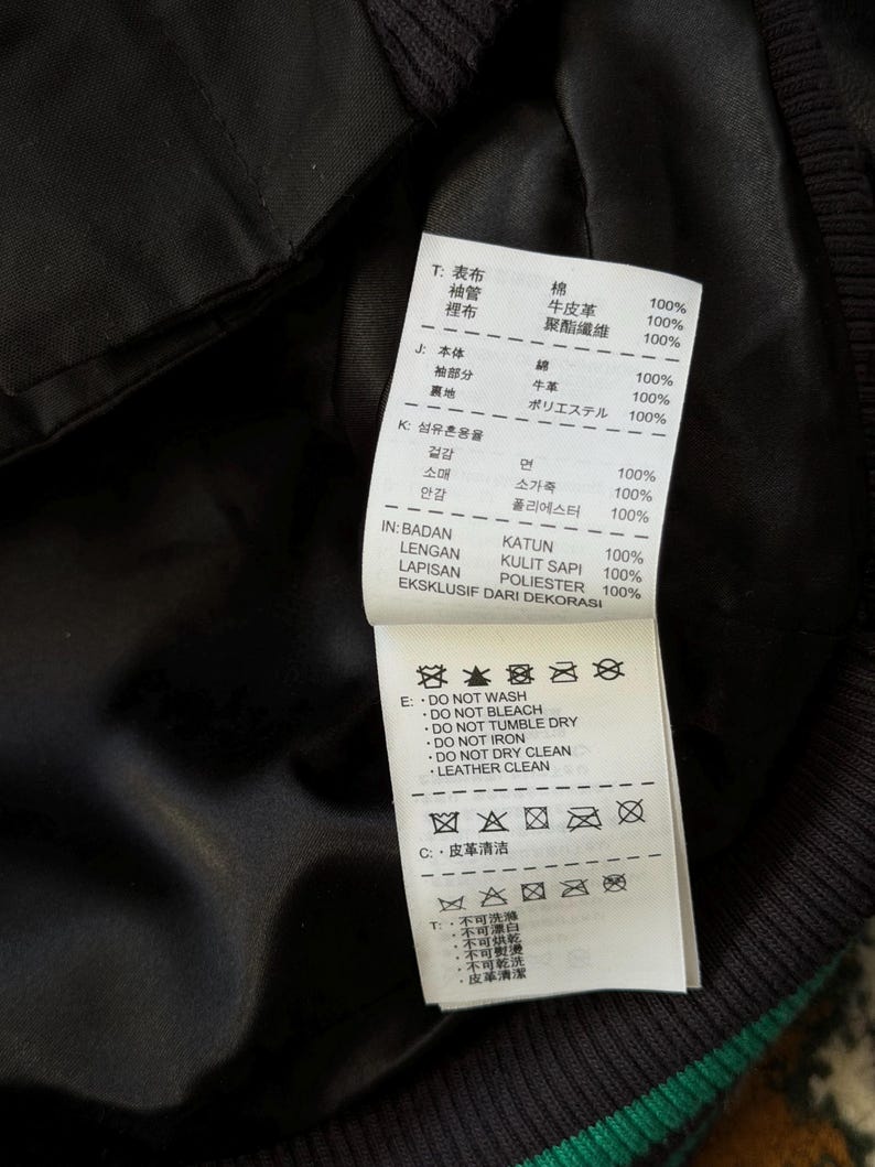 Może przedstawiać: Zbliżenie na czarną podszewkę kurtki z białą metką. Metka zawiera tekst w wielu językach, w tym japońskim i angielskim, szczeg&oacute;łowo opisujący skład tkaniny i instrukcje pielęgnacji. Kurtka ma zielony pasek.