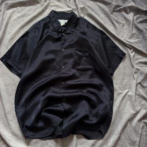 May include: A black short-sleeved button-down shirt with a subtle polka dot pattern. The shirt has a chest pocket and a classic collar. The fabric appears to be a smooth, possibly silk-like material, with a slight sheen. The shirt is laid flat on a gray surface.