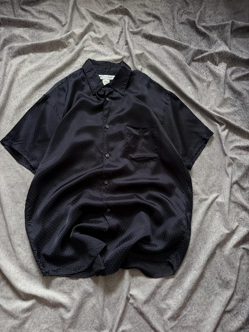 May include: A black short-sleeved button-down shirt with a subtle polka dot pattern. The shirt has a chest pocket and a classic collar. The fabric appears to be a smooth, possibly silk-like material, with a slight sheen. The shirt is laid flat on a gray surface.