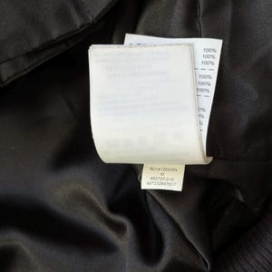 Może przedstawiać: Zbliżenie na wnętrze czarnego ubrania, ukazujące białą metkę z tekstem i procentami. Etykieta zawiera tekst "SU141203GIN M 650727-010 887232947637" oraz procenty, takie jak "100%" i "0%."