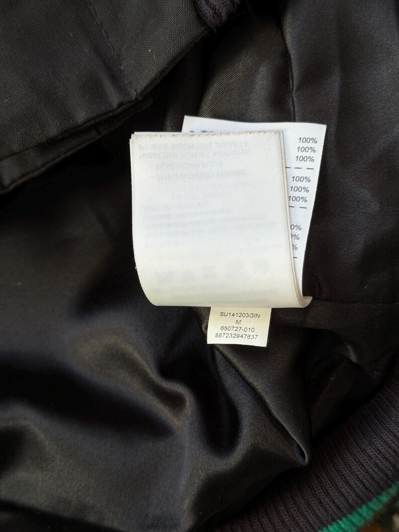 Może przedstawiać: Zbliżenie na wnętrze czarnego ubrania, ukazujące białą metkę z tekstem i procentami. Etykieta zawiera tekst "SU141203GIN M 650727-010 887232947637" oraz procenty, takie jak "100%" i "0%."