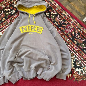 May include: A gray Nike hoodie with a yellow-lined hood and a large yellow Nike logo across the chest. The hoodie has a kangaroo pocket and yellow drawstrings. The garment is made of a soft material.