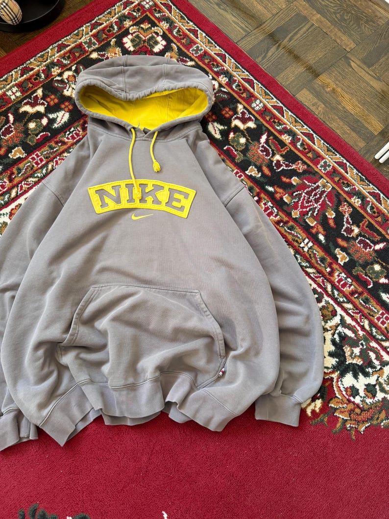 May include: A gray Nike hoodie with a yellow-lined hood and a large yellow Nike logo across the chest. The hoodie has a kangaroo pocket and yellow drawstrings. The garment is made of a soft material.