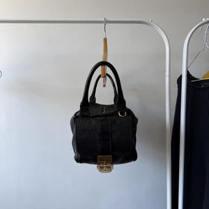 May include: Black leather handbag with a textured finish, gold-tone hardware, and a top handle. The bag is hanging on a wooden hanger from a white metal clothing rack. The bag has a flap closure with a gold clasp.