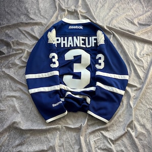 May include: A blue hockey jersey with white accents and the name "PHANEUF" and the number "3" in white. The jersey has the Reebok logo and maple leaf details on the shoulders. The jersey is laid flat on a gray surface.