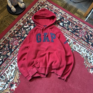 Puede incluir: Una sudadera con capucha de forro polar rojo que presenta la palabra GAP en letras texturizadas de color azul oscuro en el pecho. La prenda incluye un bolsillo tipo canguro frontal, mangas largas con puños acanalados y un cordón rojo.