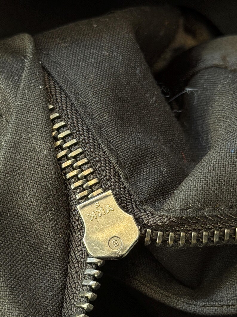 Może przedstawiać: Zbliżenie czarnej tkaniny ze srebrnym zamkiem błyskawicznym. Zamek jest częściowo zapięty, odsłaniając zęby i logo marki YKK na suwaku. Tkanina wydaje się być wytrzymałym materiałem.