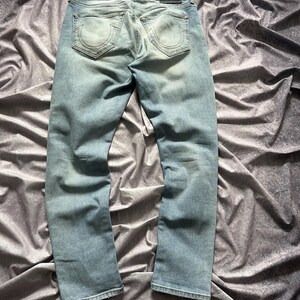 May include: Light blue denim jeans with a faded wash. The jeans feature a classic five-pocket design, a leather patch, and a straight leg. The back pockets have a distinctive horseshoe-shaped stitching. The jeans are laid flat on a textured, gray surface.