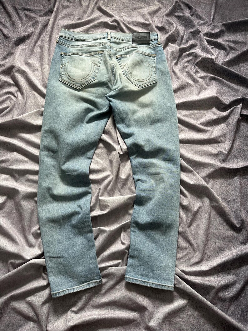 May include: Light blue denim jeans with a faded wash. The jeans feature a classic five-pocket design, a leather patch, and a straight leg. The back pockets have a distinctive horseshoe-shaped stitching. The jeans are laid flat on a textured, gray surface.