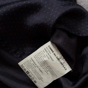 May include: Close-up of a navy blue fabric with small blue polka dots. A white tag with black text is visible, indicating the garment is made in France by Comme des Gar&ccedil;ons. The tag provides care instructions.