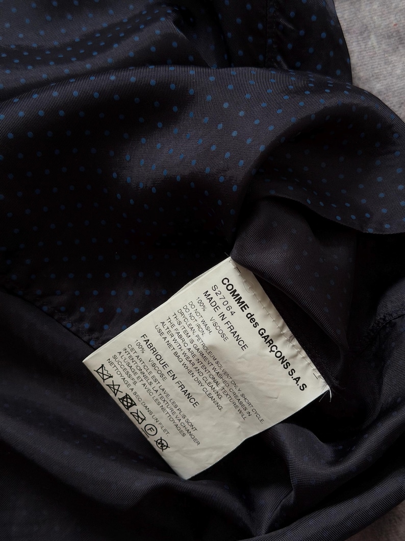 May include: Close-up of a navy blue fabric with small blue polka dots. A white tag with black text is visible, indicating the garment is made in France by Comme des Gar&ccedil;ons. The tag provides care instructions.