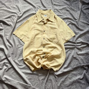 Puede incluir: Una camisa de manga corta con botones, de color amarillo claro con rayas verticales marrones. La camisa tiene cuello, bolsillo en el pecho y las letras "C.P." en el cuello. La camisa es de tejido ligero.