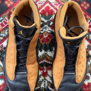 Op de afbeelding: Paar bruin en zwarte Air Jordan 13 sneakers. De schoenen hebben een bruin bovenwerk met zwarte accenten, zwarte veters en het woord "JORDAN" op de neus. Het Jumpman-logo is zichtbaar op de tong.