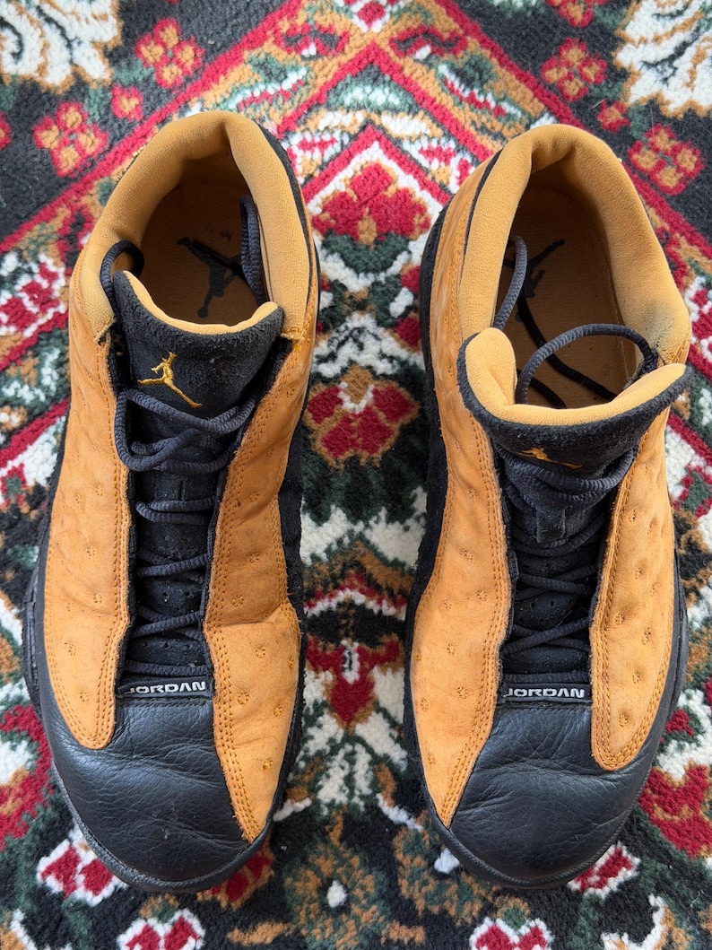 Op de afbeelding: Paar bruin en zwarte Air Jordan 13 sneakers. De schoenen hebben een bruin bovenwerk met zwarte accenten, zwarte veters en het woord "JORDAN" op de neus. Het Jumpman-logo is zichtbaar op de tong.