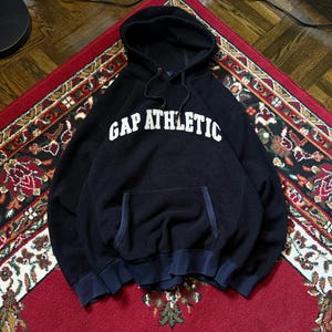 May include: A navy blue hooded sweatshirt with the text "GAP ATHLETIC" in white block letters. The sweatshirt features a front pocket and a drawstring hood. The garment is displayed on a red patterned rug.