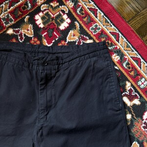 May include: Close-up of black trousers with a button fastening. The trousers are made of a solid, dark fabric. The image is taken from a high angle, showing the waist and upper portion of the trousers.