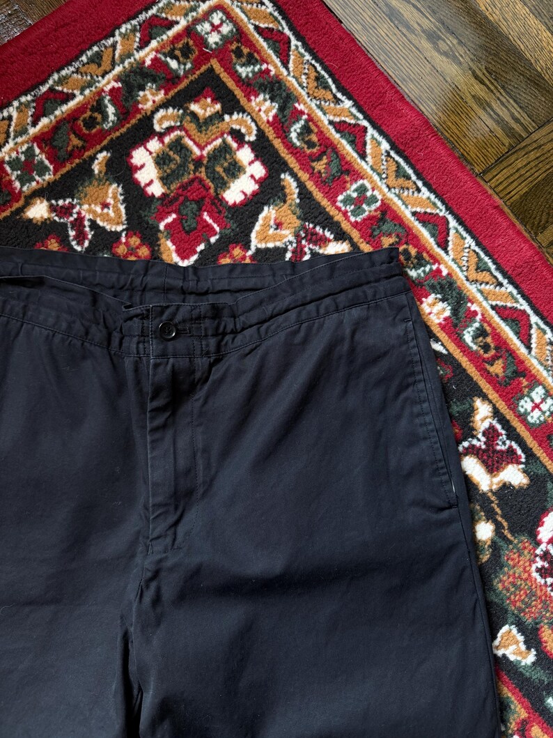 May include: Close-up of black trousers with a button fastening. The trousers are made of a solid, dark fabric. The image is taken from a high angle, showing the waist and upper portion of the trousers.