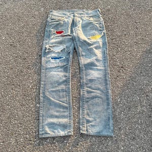 Puede incluir: Un par de pantalones vaqueros de mezclilla gris claro con varios parches de diferentes colores cosidos en la parte delantera. Los parches son rojo, amarillo, azul y blanco.