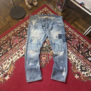 Könnte beinhalten: Hellblaue Jeans mit mehreren Patches in verschiedenen Mustern und Farben. Die Jeans sind abgenutzt und weisen sichtbare Gebrauchsspuren auf. Die Jeans liegen auf einem roten Musterteppich.