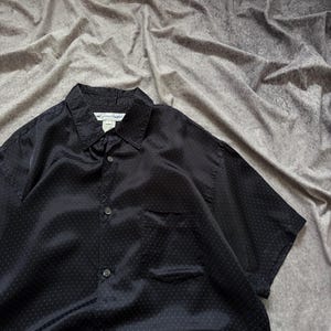 May include: Black short-sleeve button-down shirt with a subtle polka dot pattern. The shirt has a chest pocket and a collar. The tag reads "The Shirt". The fabric has a slight sheen.