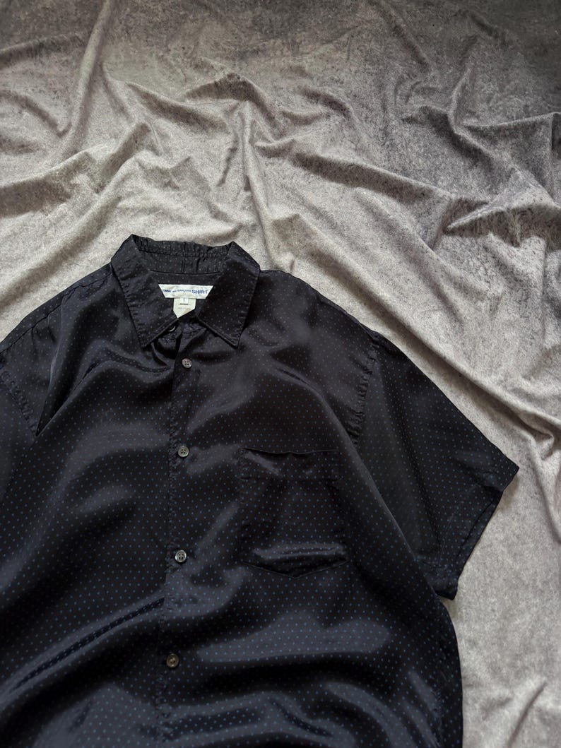 May include: Black short-sleeve button-down shirt with a subtle polka dot pattern. The shirt has a chest pocket and a collar. The tag reads "The Shirt". The fabric has a slight sheen.