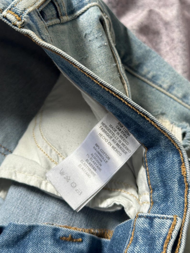 May include: Close-up of light blue denim jeans with a white tag. The tag reads "MADE IN MEXICO" and lists the fabric composition as 99% cotton and 1% elastane. The jeans have visible stitching and a worn appearance.