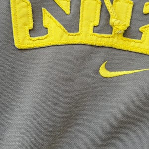 May include: Gray hoodie with yellow embroidered "NYC" lettering and a yellow Nike swoosh. The hoodie features a drawstring and a textured fabric. The yellow lettering and swoosh provide a striking contrast against the gray background.