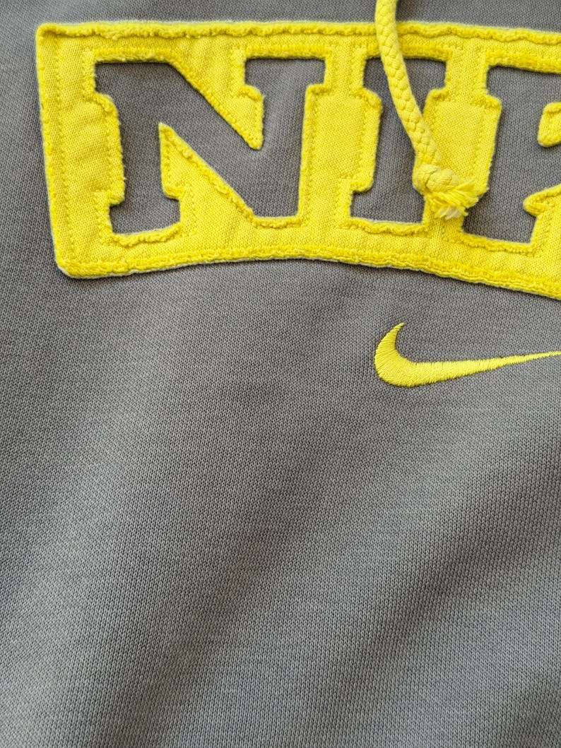 May include: Gray hoodie with yellow embroidered "NYC" lettering and a yellow Nike swoosh. The hoodie features a drawstring and a textured fabric. The yellow lettering and swoosh provide a striking contrast against the gray background.