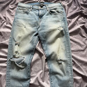 May include: Light blue denim jeans with distressed details, including rips and fading. The jeans have a classic five-pocket design and a button and zipper closure. The fabric appears to be a medium weight.