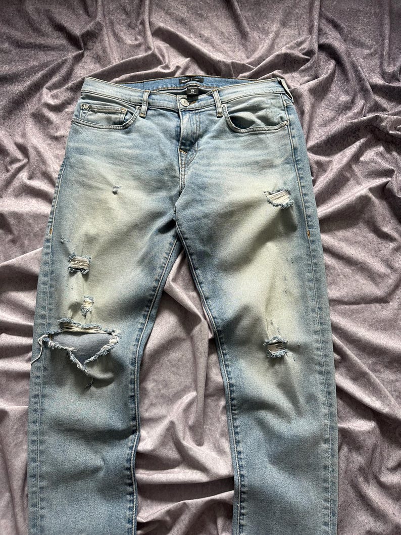 May include: Light blue denim jeans with distressed details, including rips and fading. The jeans have a classic five-pocket design and a button and zipper closure. The fabric appears to be a medium weight.