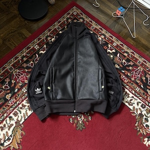 May include: Black zip-up track jacket with a faux leather front panel. The jacket features black stripes on the sleeves, the Adidas logo, and a ribbed collar and cuffs. The jacket is laid on a red patterned rug.