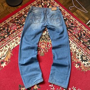 May include: Light blue denim jeans with a faded wash, visible back pockets, and a leather patch. The jeans are laid out on a red patterned rug, showcasing the back view of the garment.