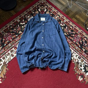Puede incluir: Camisa vaquera azul medio de manga larga, abotonada y con un bolsillo en el pecho. La prenda está colocada sobre una alfombra roja y negra estampada.