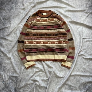 May include: A brown, beige and pink striped sweater with a geometric pattern and small deer silhouettes. The sweater has a crew neck and long sleeves.